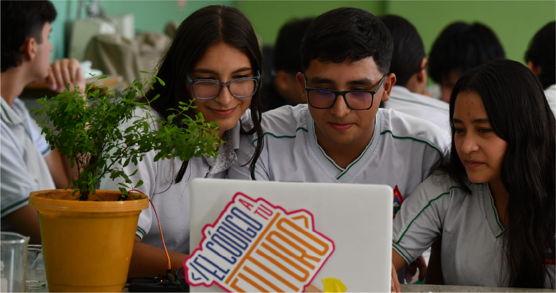 Tres estudiantes con uniforme escolar trabajan juntos frente a un portátil, mientras observan un experimento con una planta conectada a un dispositivo electrónico en un aula.
