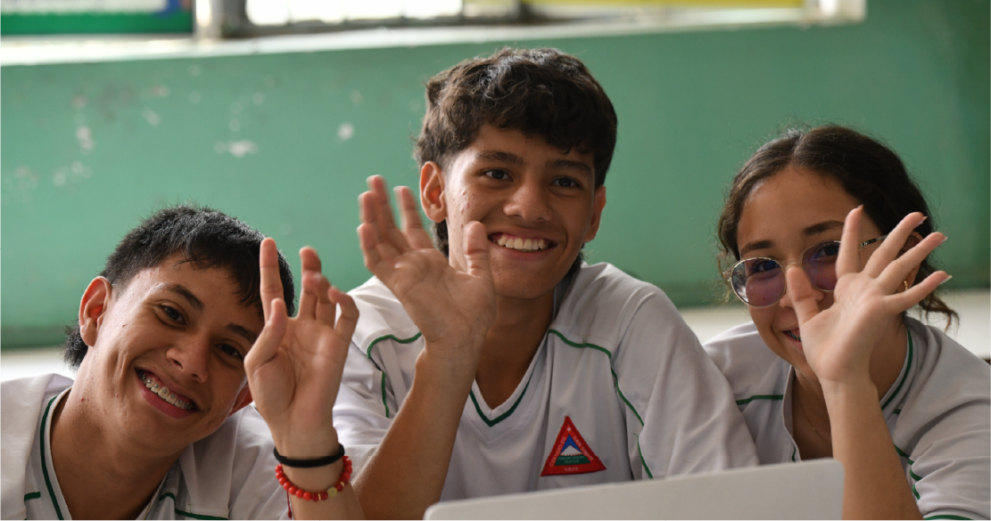 Tres estudiantes con uniforme escolar sonríen, mientras están sentados frente a un portátil en un aula.
