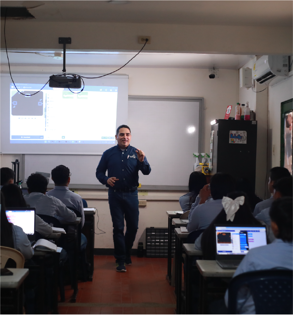 hombre dictando clase en un aula con la proyección de una pantalla a sus espaldas.