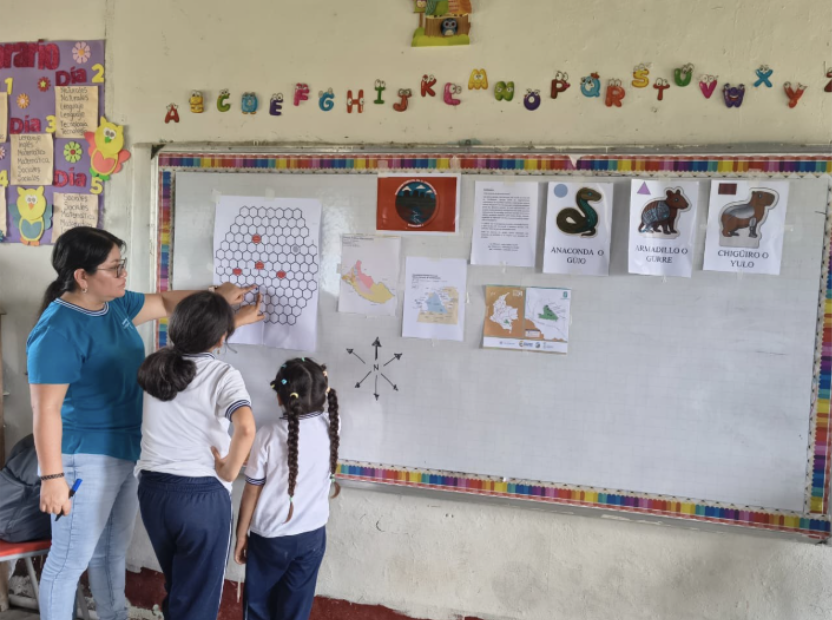 Docente y estudiantes observan materiales educativos expuestos en la pared de un aula.