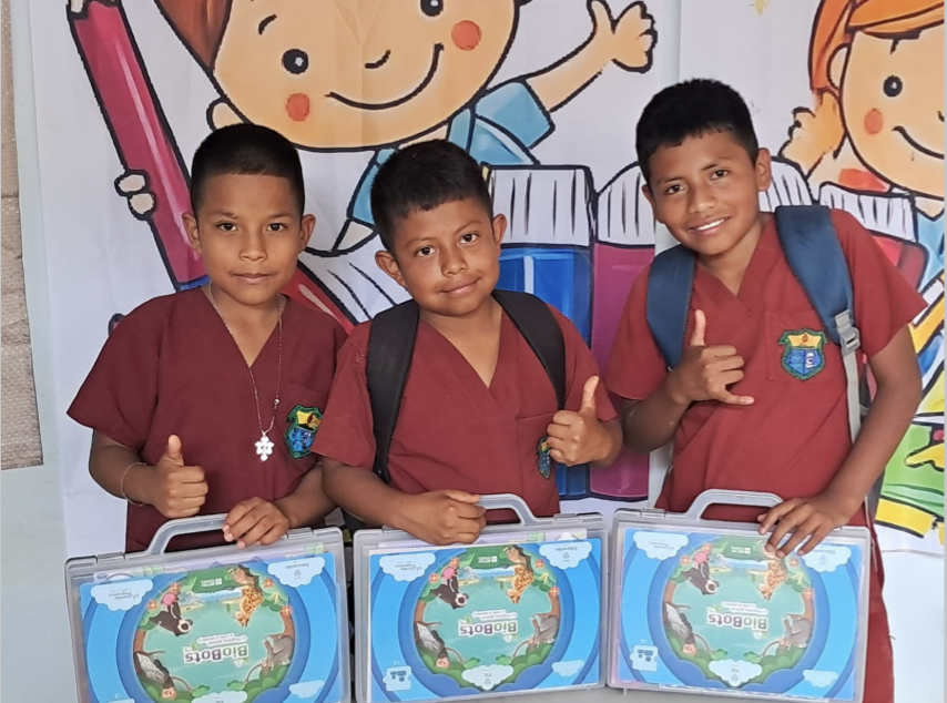 Tres estudiantes con uniforme escolar sostienen kits educativos de Biobots y hacen gestos con las manos frente a un mural infantil.