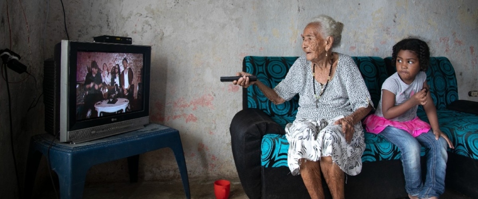 Foto de un adulto mayor junto a una menor de edad viendo televisión