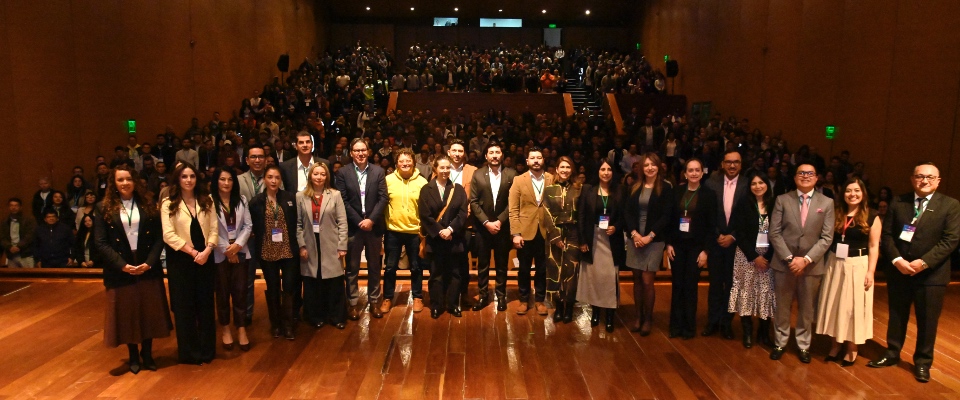Grupo de profesionales posando en un auditorio con escenario de madera, frente a un público numeroso. Visten trajes formales y credenciales, en lanzamiento de AVANZATEC 2026 con iluminación cálida.