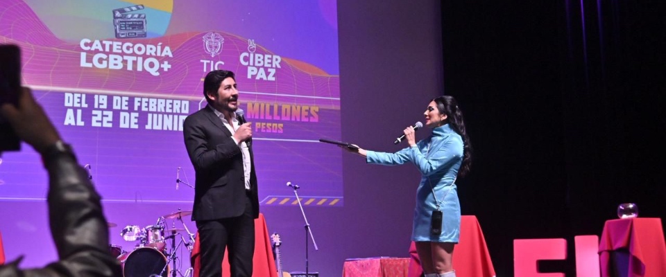 En un escenario iluminado, una presentadora interactua con el vicemninistro frente a una pantalla que anuncia la categoría LGBTIQ+ y fechas del evento. Al fondo se ven instrumentos y mesas rojas.