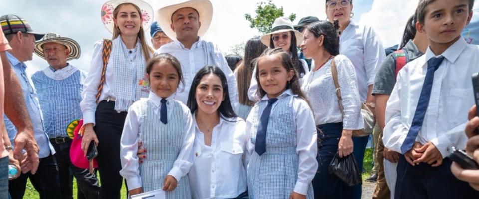 Niñas con uniforme escolar sonríen junto a Ministra TIC en acto comunitario rural, rodeadas de asistentes y autoridades bajo cielo despejado.
