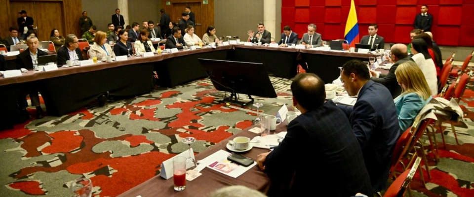 Funcionarios y asistentes participan en reunión oficial alrededor de mesa en U, con bandera de Colombia y pantallas al centro