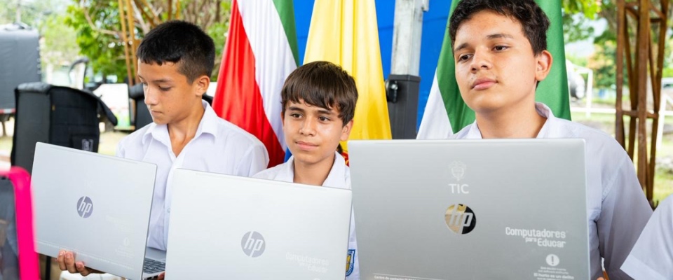 Tres estudiantes con uniforme sostienen portátiles del programa Computadores para Educar frente a banderas, durante entrega de equipos TIC