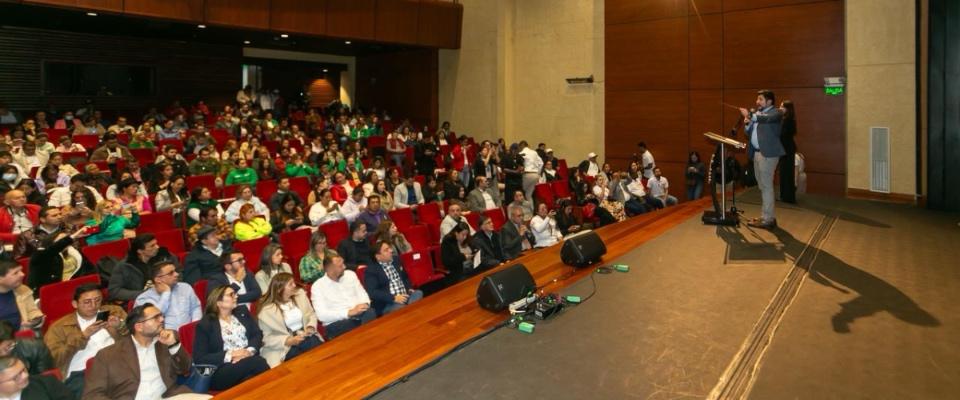 Conferencista interviene desde el escenario ante auditorio lleno durante evento institucional con participación de ciudadanos y expertos