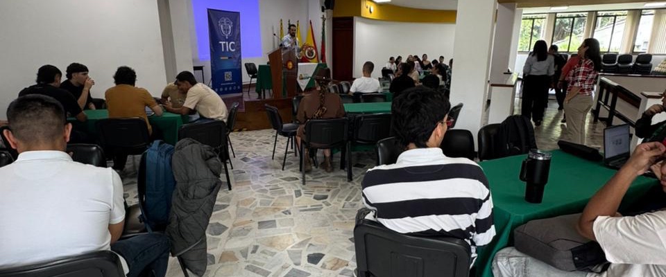 En un salón de Pereira, personas sentadas en mesas escuchan a un expositor frente a un podio del TIC, en encuentro sobre Educación Digital