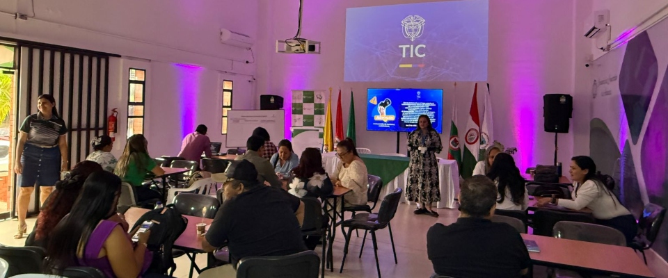 Docentes y participantes dialogan en mesas de trabajo mientras una moderadora dirige una sesión sobre educación digital en un salón institucional.