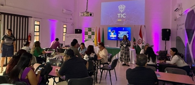 Docentes y participantes dialogan en mesas de trabajo mientras una moderadora dirige una sesión sobre educación digital en un salón institucional.