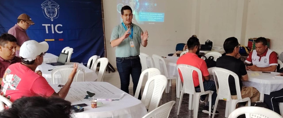 En un salón comunitario en Mitú, un facilitador expone frente a varias mesas donde participantes dialogan y escriben ideas para el CONPES de Educación Digital.