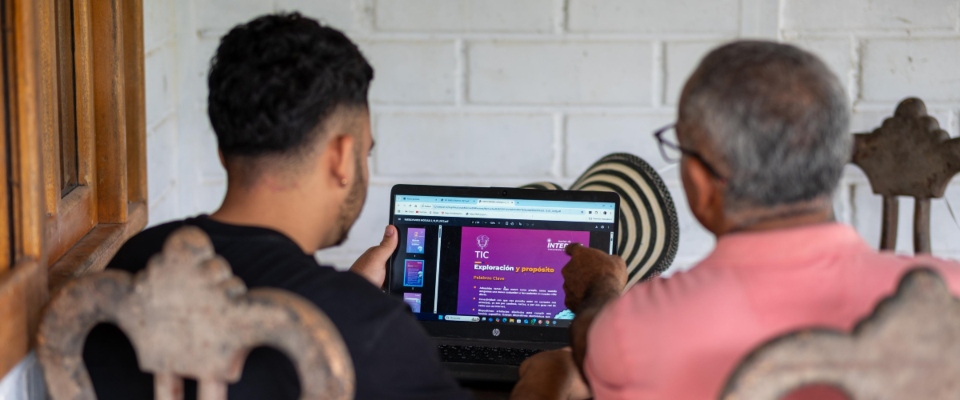 Un joven y un adulto mayor miran la pantalla de un portátil con información del sector TIC, reflejando el acceso a nuevas tecnologías en Colombia.