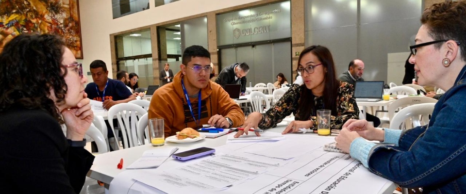 Cuatro personas dialogan en una mesa con documentos y laptops durante un taller; detrás, más grupos trabajan en construir la política de educación digital.