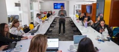 Personas trabajan con portátiles alrededor de una mesa larga en una sala moderna; un hombre de pie habla mientras el grupo participa en la discusión