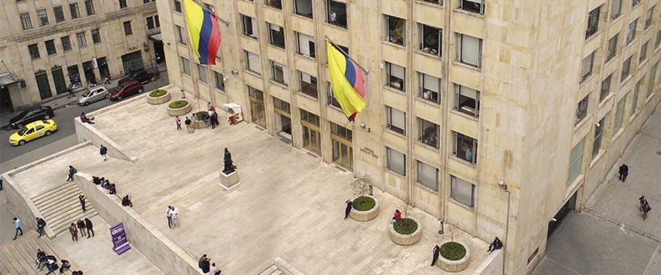 Vista aérea de edificio oficial con banderas de Colombia; personas transitan la plaza, reflejando presencia estatal y acceso institucional.