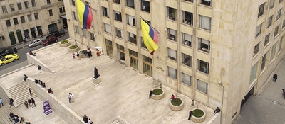 Vista aérea de edificio oficial con banderas de Colombia; personas transitan la plaza, reflejando presencia estatal y acceso institucional.