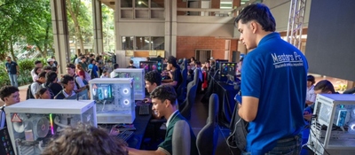 Estudiantes y jóvenes usan computadores en un evento tecnológico en campus abierto; filas de equipos y asistentes interactúan activamente.