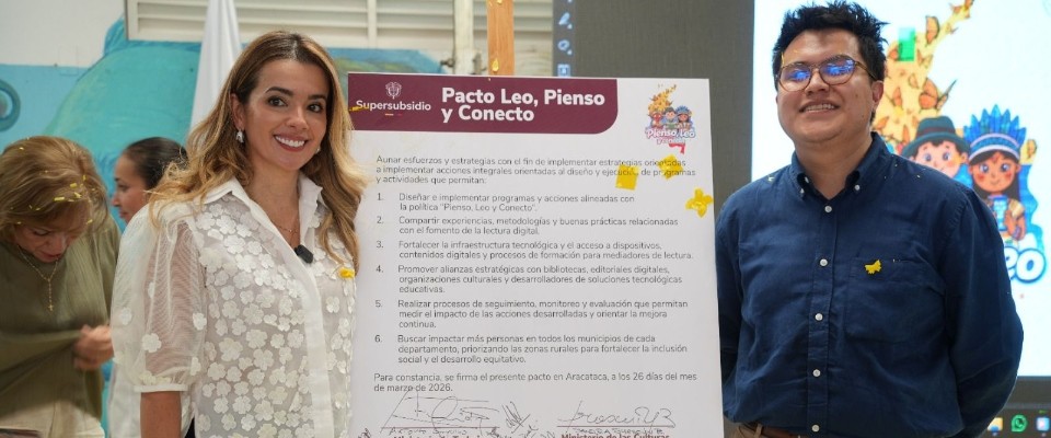 Hombre y mujer posan con afiche oficial del pacto de lectura digital. Al fondo, niños y dibujos que reflejan entorno escolar