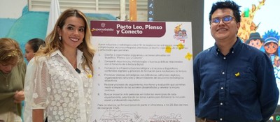Hombre y mujer posan con afiche oficial del pacto de lectura digital. Al fondo, niños y dibujos que reflejan entorno escolar