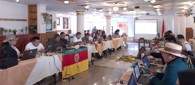 Personas indígenas y funcionarios trabajan con portátiles en mesa larga, en reunión sobre derechos comunicativos liderada por MinTIC