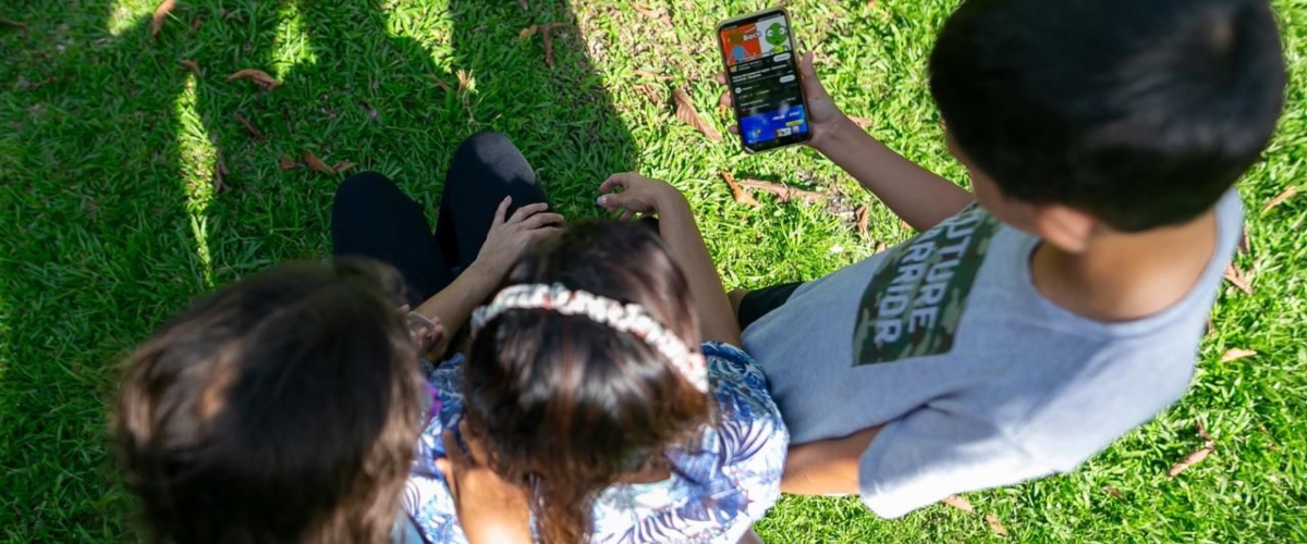 Niños comparten un teléfono en el pasto, combinando juego y tecnología en un momento de ocio al aire libre.