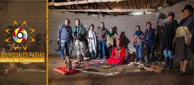 Grupo diverso en vivienda rural indígena; personas con cámaras rodean a mujer en traje tradicional y elementos rituales en el suelo.