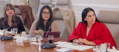 Tres mujeres en mesa de reunión oficial escuchan atentas; hay micrófonos y documentos, en un entorno institucional.