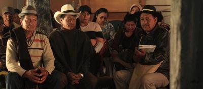 Grupo de hombres y mujeres indígenas sentados en reunión comunitaria; uno habla con cuaderno en mano mientras otros escuchan atentos.