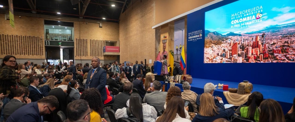 Auditorio lleno en evento oficial; en tarima, pantalla muestra ciudad y lema “Colombia, el país de la belleza” ante asistentes atentos.