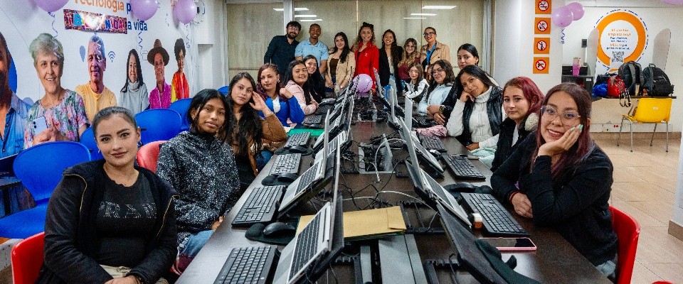 Grupo de mujeres en aula TIC con computadores, sonríen a cámara durante formación técnica impulsada por MinTIC e IPES.