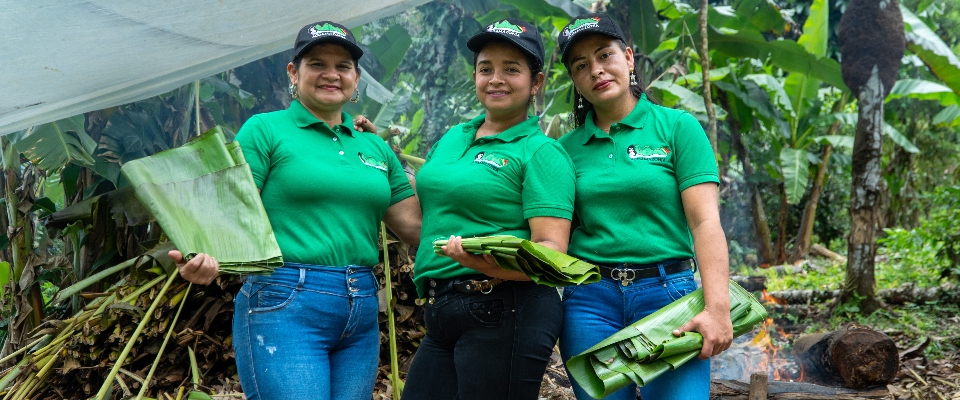 Tres mujeres campesinas con camisetas verdes sostienen hojas de plátano en entorno rural; representan lideresas de “Guardianas de Tierra Viva”