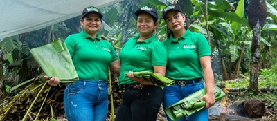 Tres mujeres campesinas con camisetas verdes sostienen hojas de plátano en entorno rural; representan lideresas de “Guardianas de Tierra Viva”