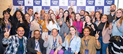 Grupo diverso de personas sonríe frente a fondo “CiberPaz TIC”, levantando signos de paz; ambiente colaborativo por la inclusión digital