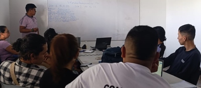 Grupo de personas en aula escucha a un expositor frente a tablero; escena educativa que evoca memoria y arraigo territorial