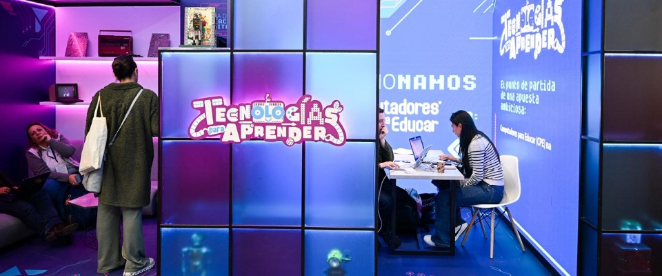 Stand iluminado “Tecnologías para Aprender” en feria; personas exploran y trabajan con laptops. TIC promueve uso responsable e innovación