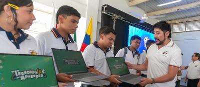 Estudiantes con uniforme reciben portátiles de un instructor; al fondo, bandera de Colombia y pantalla, impulso digital en Sucre