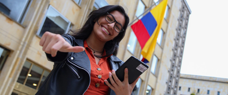 Joven sonriente con gafas señala al frente y sostiene tablet; al fondo bandera de Colombia y edificio institucional del Ministerio TIC