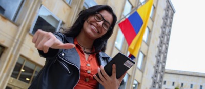 Joven sonriente con gafas señala al frente y sostiene tablet; al fondo bandera de Colombia y edificio institucional del Ministerio TIC