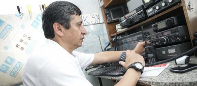 Hombre opera radios y equipos de comunicación en una mesa técnica, ajustando perillas y un walkie-talkie en entorno de radioafición.