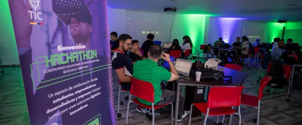 Personas trabajan en laptops durante hackathon TIC; mesas con equipos y luces verdes. Evento Colombia 5.0 reúne expertos y talleres