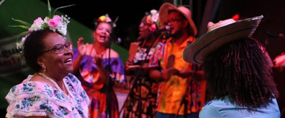 Mujer sonriente con flores en el cabello disfruta música en vivo; detrás, grupo canta y toca con vestuario colorido afro