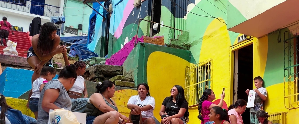 Mujeres y niños conversan en escaleras de barrio con casas coloridas; escena comunitaria ligada a líderes que transforman territorios.