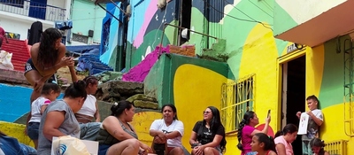 Mujeres y niños conversan en escaleras de barrio con casas coloridas; escena comunitaria ligada a líderes que transforman territorios.