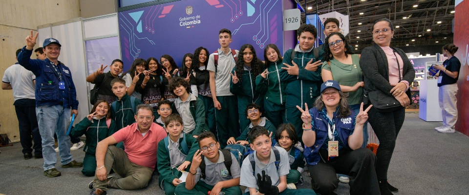 Grupo de estudiantes y adultos posan en feria con logo Gobierno de Colombia; sonrisas y gestos celebran CiberPaz y ciudadanía digital.