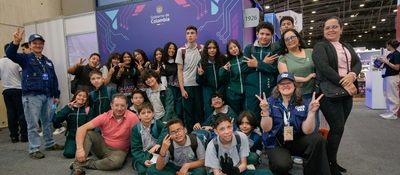 Grupo de estudiantes y adultos posan en feria con logo Gobierno de Colombia; sonrisas y gestos celebran CiberPaz y ciudadanía digital.
