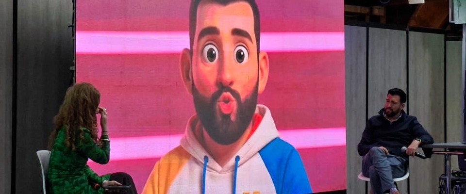 Panel sobre medios públicos y conectividad con dos personas en escenario y personaje animado proyectado en pantalla gigante.