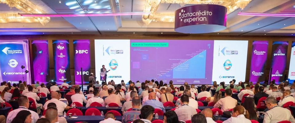 Auditorio lleno durante evento tecnológico con pantallas gigantes y asistentes atentos a charla sobre transformación digital