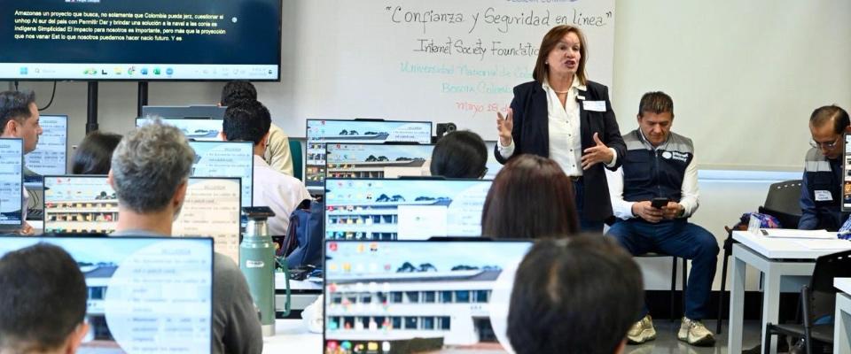 Personas trabajan en computadores durante una capacitación técnica en Bogotá, liderada por una mujer en un aula tecnológica.