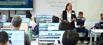 Personas trabajan en computadores durante una capacitación técnica en Bogotá, liderada por una mujer en un aula tecnológica.